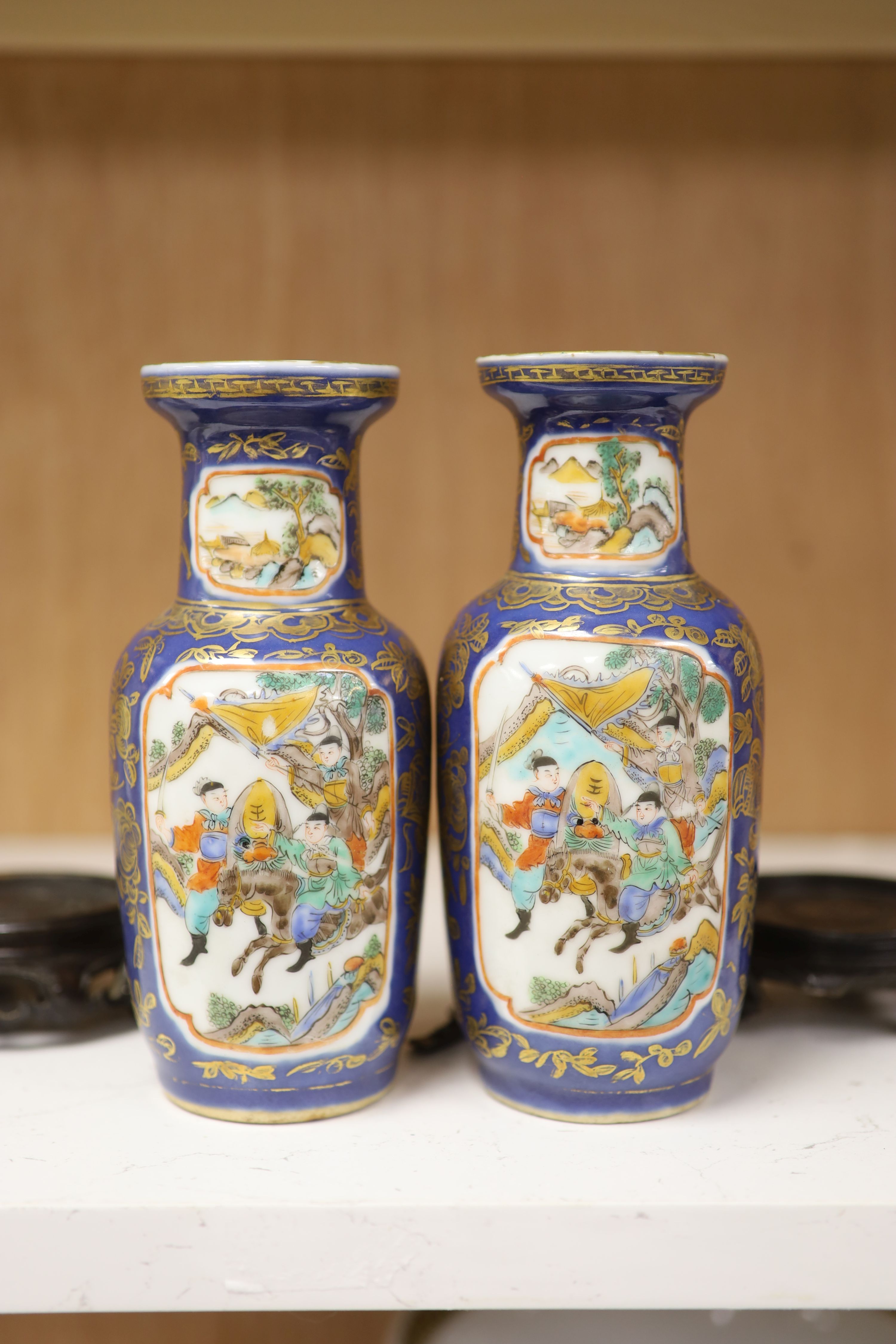 A pair of Chinese Kangxi style blue-ground vases, late 19th century decorated with panels of warriors and heightened in gilt, on carved hardwood stands, height 18cm excluding stand